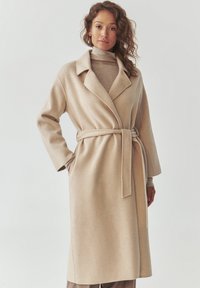 Beige kabát z vlněné směsi s šálovým límcem, dlouhými rukávy, páskem v pase a volným střihem. Hladká textura se bočními kapsami.