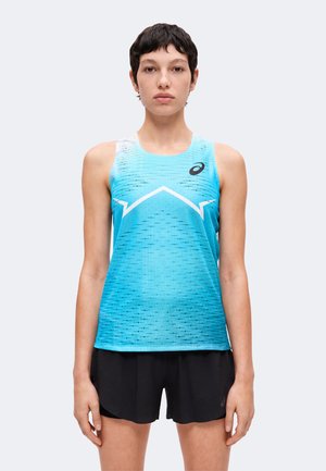 Ung person med kort mørkt hår iført en blå ærmeløs sportstop med hvidt design og sorte atletikshorts, stående mod en grå baggrund.