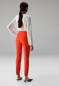 Blouse blanche à manches longues, pantalon orange ajusté, et mules noires à talons. Tissu lisse avec une silhouette élégante et épurée.