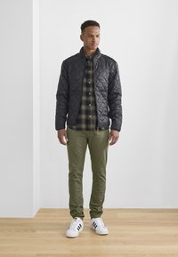 Svart quiltet jakke med høy krage, grønn rutet skjorte, olivenfargede bukser og hvite sneakers med svarte striper. Står på tregulv.