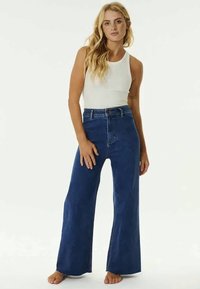 Kvinna som står barfota iklädd en vit ärmlös topp och högmidjade vida blå jeans mot en enfärgad bakgrund.