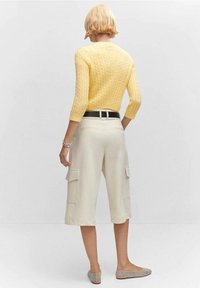 Keltainen neulottu villapaita, jossa on kolmevartiset hihat, yhdistetty kermaan sävyttyviin cargo-shortseihin, joissa on sivutaskut, sekä koristeelliset tasapohjaiset kengät.