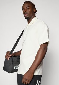 Svetlo rumena teksturirana polo majica s kratkimi rokavi in zadrgo na ovratniku, v kombinaciji z črno adidas torbico za čez ramo. Temni hlače dopolnjujejo videz.