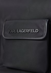 Sac à dos en tissu noir avec une poche avant dotée d'un logo noir en relief, texture lisse et couture contrastante le long des bords.