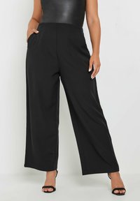 Pantalones de pierna ancha negros con cintura alta, tela suave, plisados frontales y bolsillos laterales, combinados con sandalias negras de tiras.