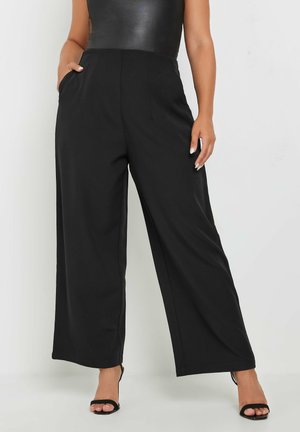 Zwarte wijde broek met hoge taille, gladde stof, plooien aan de voorkant en zijzakken, gecombineerd met zwarte sandalen met bandjes.