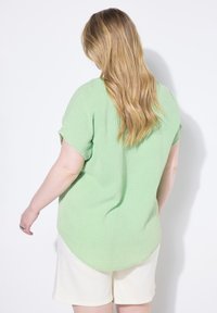 Camicetta a maniche corte a quadri verdi con orlo curvo, abbinata a pantaloni corti bianchi. Il tessuto sembra leggero e consistenza, con una vestibilità comoda.