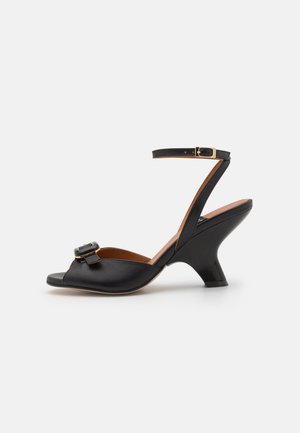 Black leather sandal with an open toe, featuring a decorative buckle and a mid-heel. Ankle strap secured with a gold-colored buckle.