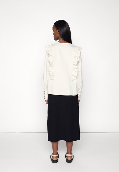 Blusa branca com acabamentos franzidos ao longo das costas, mangas longas e detalhes em botão; combinada com uma saia midi preta e sandálias.