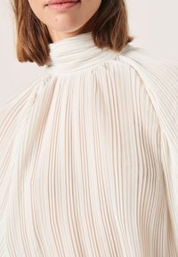 Pliss�égere blouse in een licht crèmekleurige stof met een hoge nek en een zachte, vloeiende textuur. Belangrijke kenmerken zijn diepe verticale plooien en een gestructureerde silhouet.