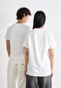 Two people standing back to back wearing plain white T-shirts, one paired with light pants, the other with dark jeans, against a white background.