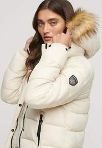 Kurtka puchowa w kolorze kremowym z kapturem wykończonym sztucznym futerkiem, zamykanymi na zamek kieszeniami oraz pikowanym wzorem. Posiada naszywkę z logo na rękawie.