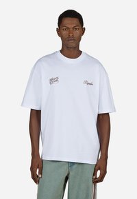 Biała bawełniana koszulka z krótkim rękawem, okrągłym dekoltem oraz haftowanym tekstem w brązowym kolorze po lewej stronie klatki piersiowej i po prawej stronie.