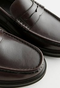Mocasines de cuero marrón oscuro con costuras decorativas, una textura lisa y una suela de goma negra de perfil bajo. Presentan un diseño recortado en la parte superior.