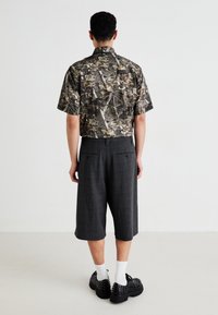 Homme portant une chemise à manches courtes avec un imprimé camouflage, un short gris à carreaux jusqu'aux genoux, des chaussettes blanches et des chaussures noires à clous, se tenant contre un mur blanc.