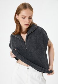 Grijze gebreide pullover met korte mouwen en een rits bij de hals. Heeft een gestructureerde stof en een geribbelde zoom, gecombineerd met witte broek.