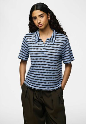 Femeie cu păr lung și creț, purtând un tricou polo tricotat cu mâneci scurte, în dungi albastre și negre, și pantaloni încrețiți în nuanțe închise, mâinile în buzunare.