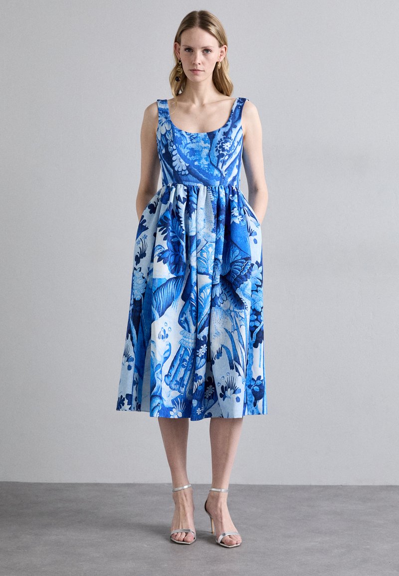Blue floral print dress with a fitted bodice, flared skirt, pockets, and scoop neckline. Styled with silver sandals.