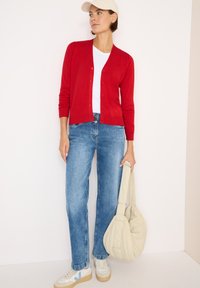 Cardigan rosso con bottoni, t-shirt bianca, jeans blu e borsa a spalla crema. Le sneakers hanno accenti chiari e scuri. Tessuti lisci.