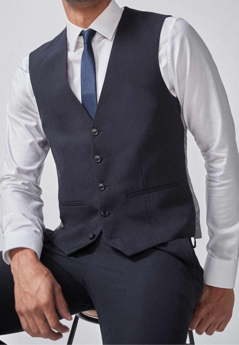 Chaleco azul marino hecho de una tela de textura suave. Presenta cinco botones, dos bolsillos frontales y un corte ajustado. Se lleva sobre una camisa blanca.