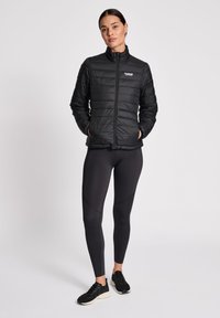 Zwarte pufferjack met horizontale stiksels en opstaande kraag, gecombineerd met zwarte legging met gebogen zoomdetails en zwarte sportschoenen.