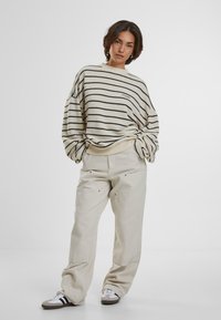 Maglione a righe in crema e blu navy con maniche ampie, abbinato a pantaloni larghi beige chiaro. Modello che indossa sneakers con accenti scuri.