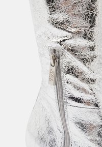 Botín metálico plateado con acabado texturizado, que cuenta con un cierre lateral y un tirador de cremallera con la marca "STEVE MADDEN."