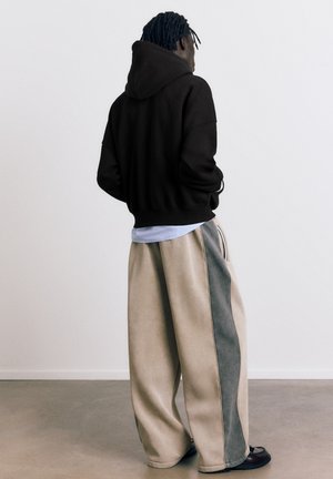 Oseba s temnimi, spletenimi lasmi, oblečena v črno jopo s kapuco, bež široke hlače s sivimi stranskimi paneli in temne čevlje, stoji na sivem tlaku.