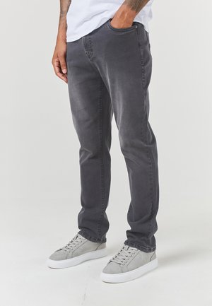 Tmavě šedé džíny se slim fit, které mají pět kapes a mírně prošlapanou texturu, v kombinaci se světle šedými teniskami na neutrálním pozadí.