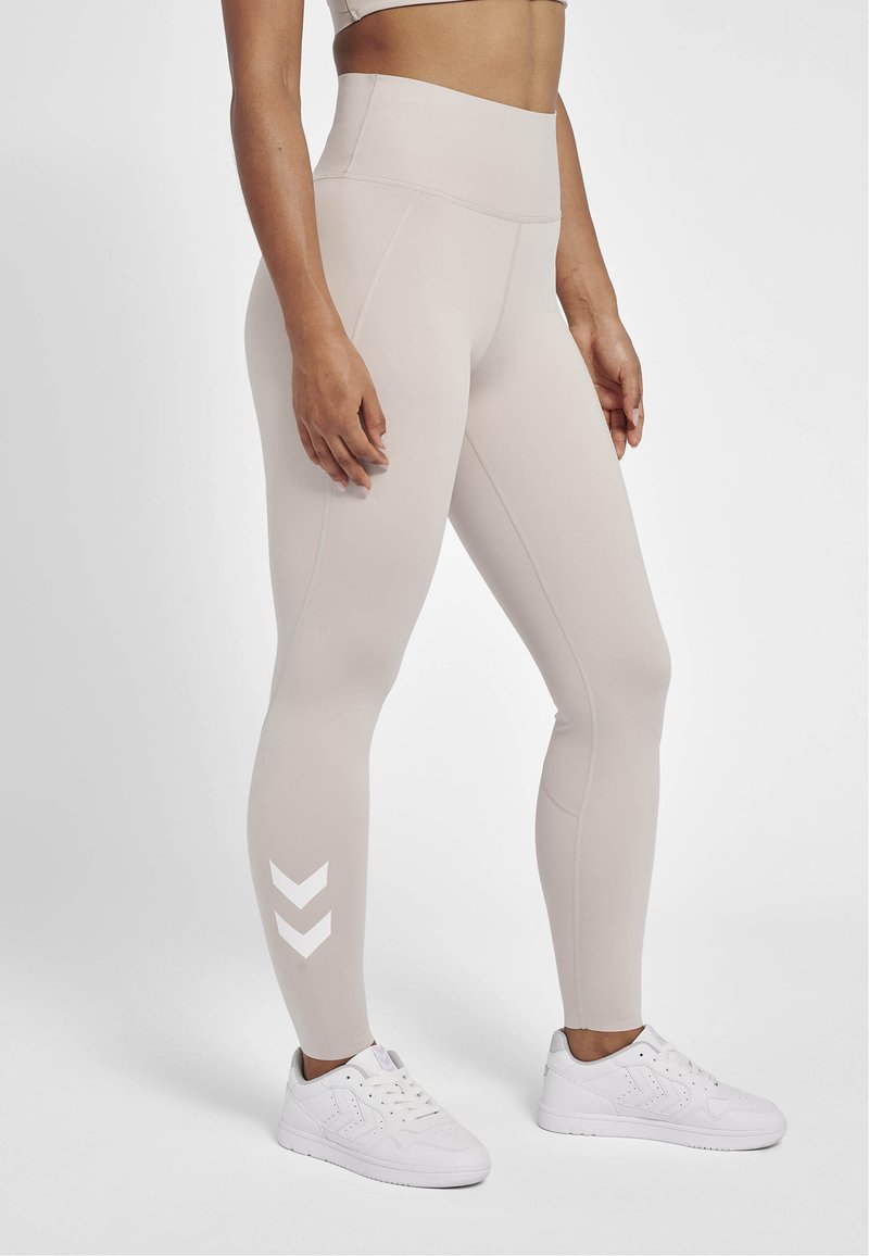 Hoge taille, beige legging met een gladde textuur en een wit zigzagpatroon op het onderste deel van het linkerpoot, gecombineerd met witte sportschoenen.