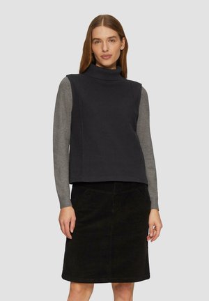 Dunkelgrauer ärmelloser Rollkragenpullover über einem hellgrauen Langarmshirt, kombiniert mit einem schwarzen Cordrock, der seitliche Taschen und eine gerade Passform hat.