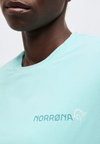 Detail osoby nosiacej svetlomodrú košeľu s logom "NORRØNA" a malou grafikou tváre s prilbou na hrudi.