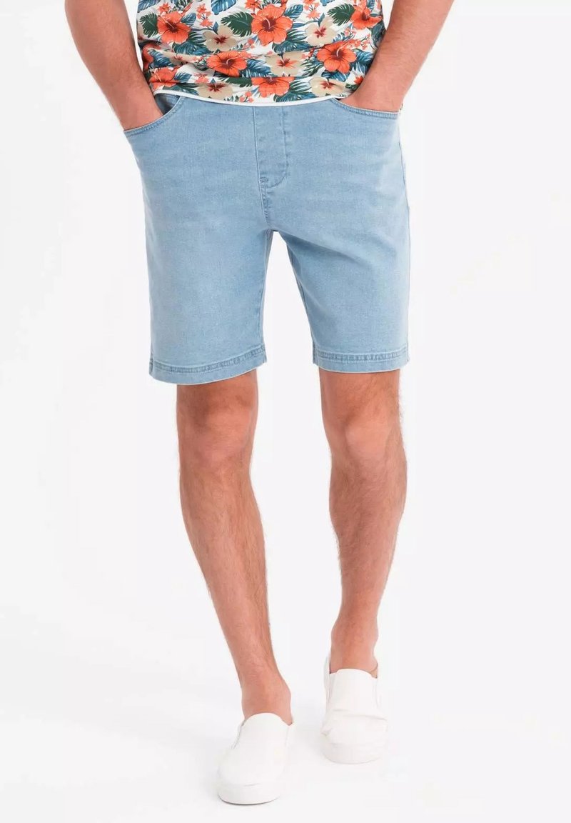 Pantaloni corti di jeans blu chiaro con due tasche laterali, taglio dritto e orlo pulito. Indossati con una camicia a stampa floreale e scarpe bianche senza lacci.