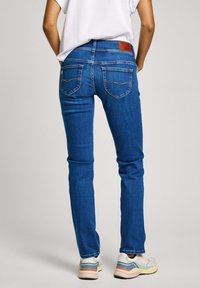 Jeans de mezclilla azul con un corte recto, que presentan una etiqueta de cuero en la cintura y costuras sutiles en los bolsillos traseros. Combinados con zapatillas.