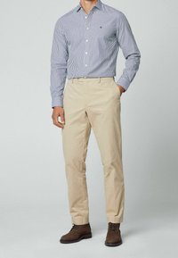 Uomo che indossa una camicia a righe verticali blu e bianche, pantaloni beigi e stivali in suede marroni con lacci, mentre sta in piedi con una mano in tasca.