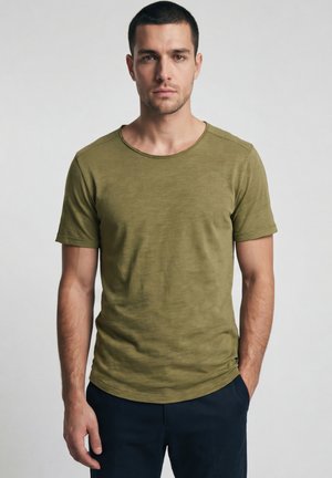 Mann mit kurzen dunklen Haaren, der ein olivgrünes kurzärmliges T-Shirt und dunkle Hosen trägt, steht vor einem schlichten hellgrauen Hintergrund.