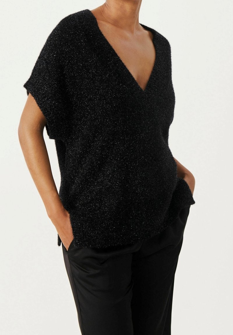 Pull noir à manches courtes avec col en V, coupe décontractée et texture brillante. Il présente des fentes latérales et des poches à l'avant. Porté avec un pantalon noir.