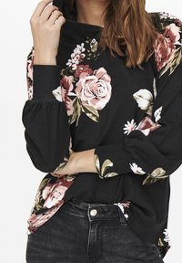 Sweat-shirt noir à fleurs avec des roses roses et blanches, manches ballon et coupe décontractée, assorti à un jean foncé.