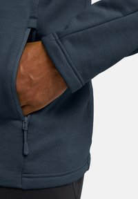 Felpa grigia scura con tasca con zip e polsini a costine. Il tessuto appare liscio con una leggera texture, progettato per comfort e funzionalità.