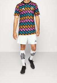 Flerfärgad vågformad mönstrad idrottströja i teal, rosa, orange och svart; kombinerad med vita shorts och svarta fotbollsskor.