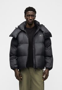 Άνδρας που φοράει μαύρο μπουφάν με φουσκωτό ύφασμα και ψηλό γιακά, μαύρο πουκάμισο και χακί παντελόνι, στέκεται μπροστά σε ένα απλό λευκό φόντο.