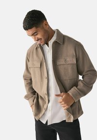 Mies, joka on pukeutunut vaaleanbeigeen nappikauluspaitaan valkoisen paidan päällä, pitäen yhtä kättään taskussa ja toista sovittaen takin etuosaa.