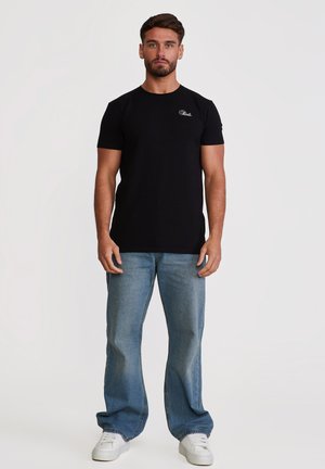 Muž stojí čelem vpřed, má na sobě černé tričko s krátkým rukávem a malým bílým logem, volné modré džíny a bílé tenisky.