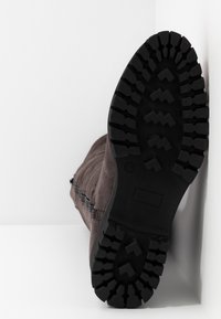 Semelle noire robuste d'une botte grise positionnée verticalement contre une surface d'angle blanche, montrant des motifs de bande de roulement profonds et texturés.