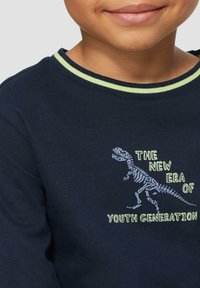 Dunkelblaues Langarmhemd aus Baumwolle, mit einem Dinosaurier-Skelett-Design und Text in Hellgrün und Gelb auf der Vorderseite.