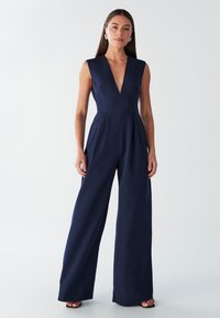 Temno modr kombinezon z globokim V-izrezom, brezrokavnikom, oprijetim pasom in širokimi hlačnicami. Gladek material z nabrani poudarki.