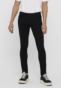 Jeans negros ajustados hechos de mezclilla, con bolsillos frontales, trabillas para el cinturón y cierre de botón. Combinados con zapatillas altas negras.