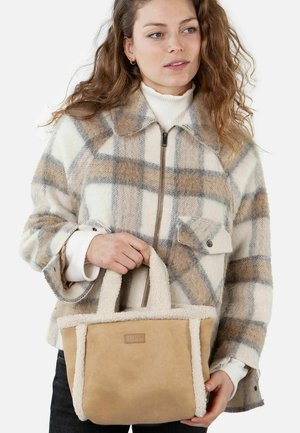 Beige suede tote bag with plush, cream-colored faux fur trim and short handles. Model wears a checkered wool jacket and white turtleneck.