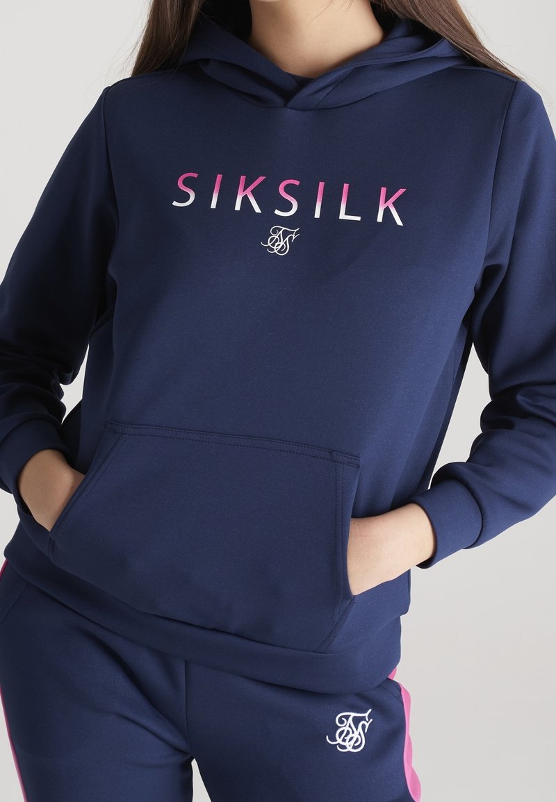Personne portant un sweat à capuche bleu marine et un pantalon assorti, le sweat porte le texte "SIKSILK" en rose et blanc, les mains dans la poche avant.