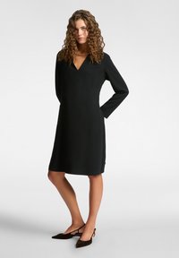 Schwarzes Langarmkleid mit V-Ausschnitt, ausgestattet mit einer Seitentasche und geradem Schnitt. Getragen mit dunkelbraunen spitzen Schuhen.